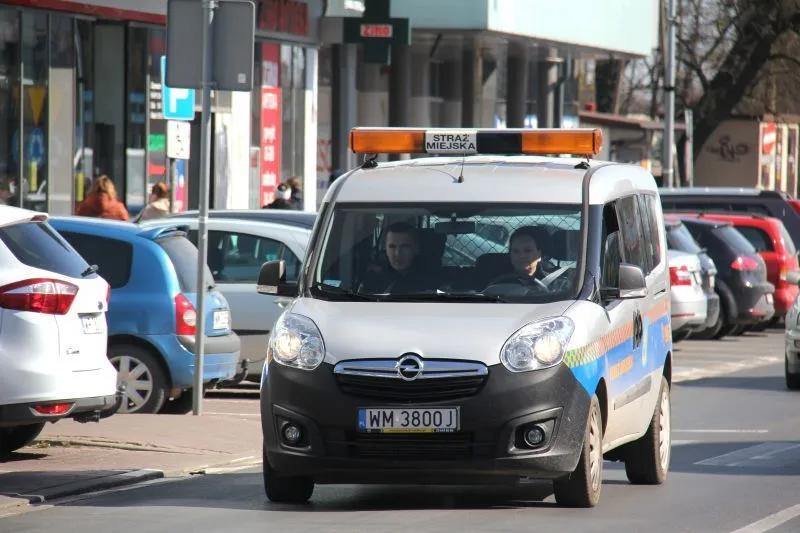 Pojazd Straży Miejskiej marki Opel z włączonymi światłami i dwoma osobami w kabinie poruszający się po miejskiej ulicy.